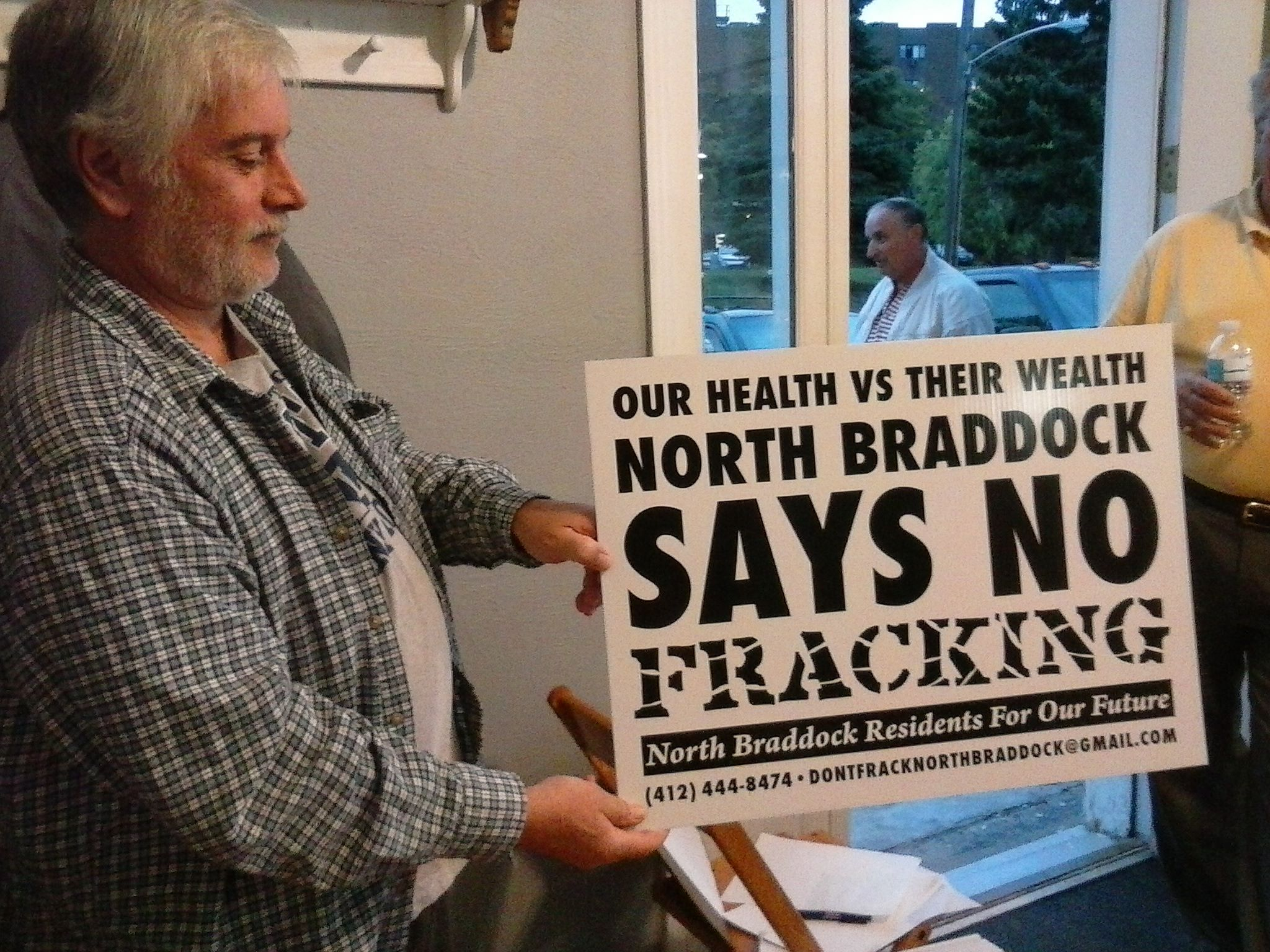 Person holding a sign produced by NBRFOF that reads "Our Health vs Their Wealth: North Braddock Says No Fracking" printed with a grant from the Mountain Watershed Association