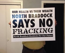 Person with obscured face holding a sign produced by NBRFOF that reads "Our Health vs Their Wealth: North Braddock Says No Fracking" printed with a grant from the Mountain Watershed Association
